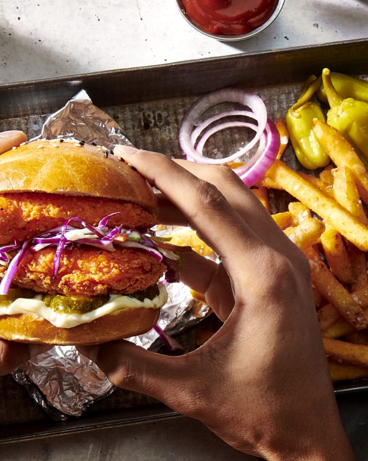 Close in view of a top down view of hands holding a chicken sandwich with a side of fries on a metal platter